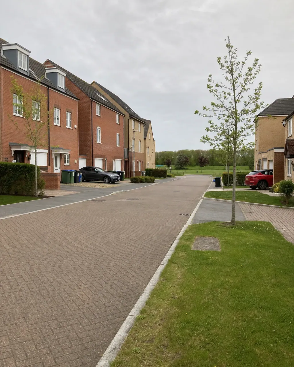 A modern Broughton estate street of brick and render houses with block-paved driveways, wide grass verges, young street trees and an open green beyond