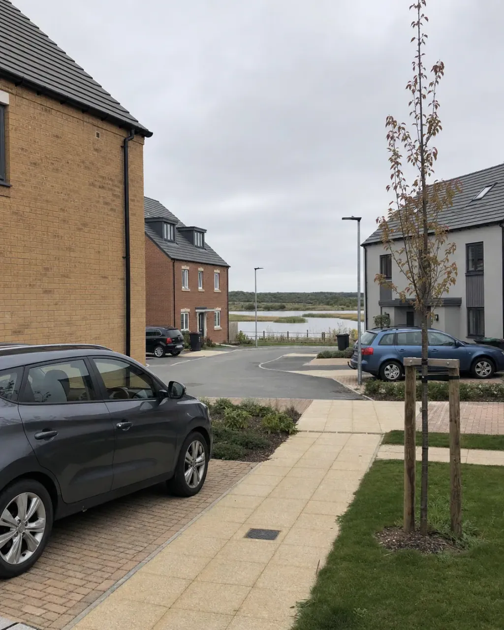 A very recent Newton Leys development of brick and grey-render houses with new block-paved driveways and young staked trees, the balancing lake visible beyond