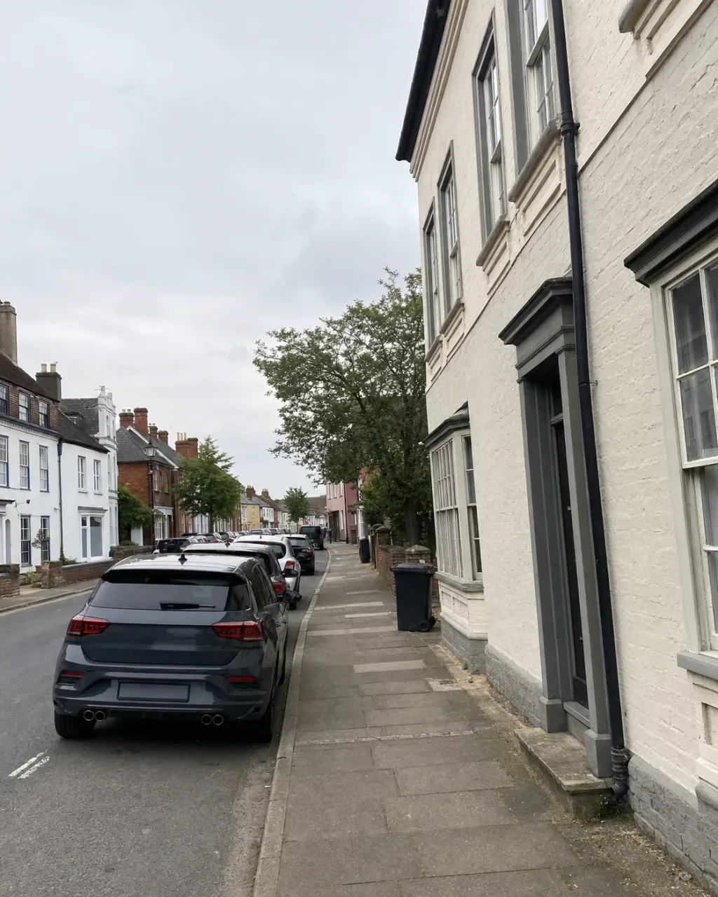 The historic Stony Stratford high street, lined with Georgian buildings in painted stucco and brick above ground-floor units, with cars parked along one side