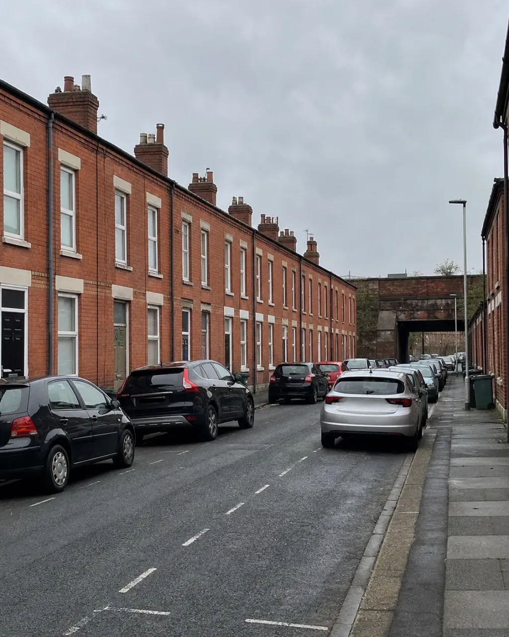 A long uniform terrace of modest red brick Victorian railway-town houses in Wolverton, opening straight onto the pavement with cars parked along both sides