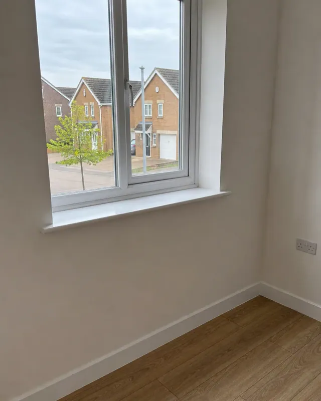 A clean bare uPVC window sill in an empty Milton Keynes new build, with laminate flooring below and newer brick housing visible through the glass