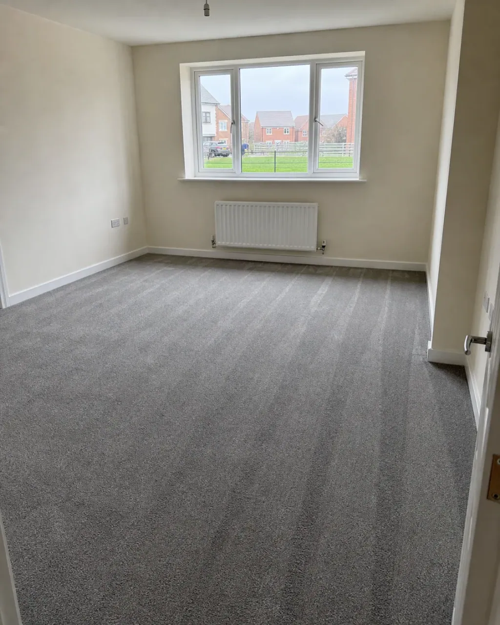 An empty Milton Keynes living room with fresh hoover lines across plain grey carpet, a large uPVC window and a radiator beneath it