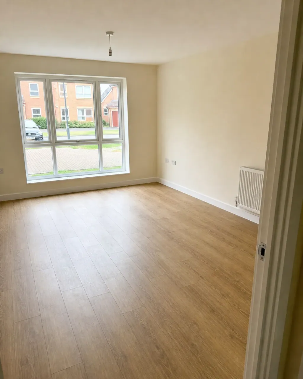 An empty Milton Keynes living room with wood-effect laminate flooring, plain magnolia walls and a large window looking onto the estate