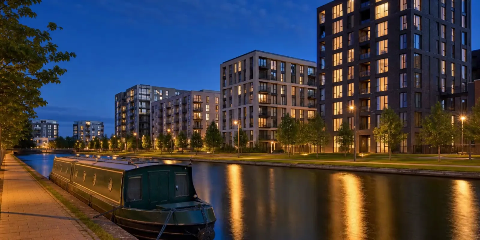 Modern canalside apartment buildings with lit windows at dusk, a narrowboat moored on the towpath in the foreground